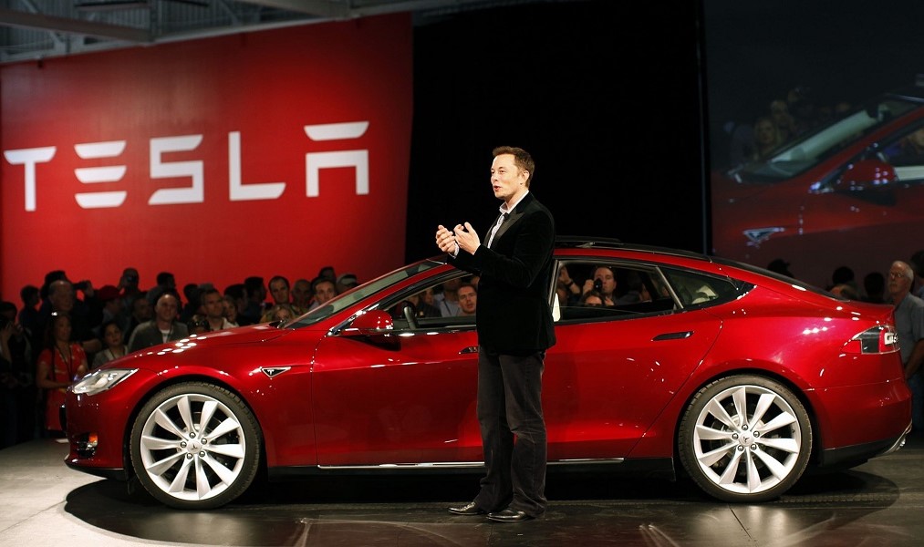Elon Musk, de roupa esporte fino, em pé ao lado de carro vermelho da Tesla durante evento da montadora