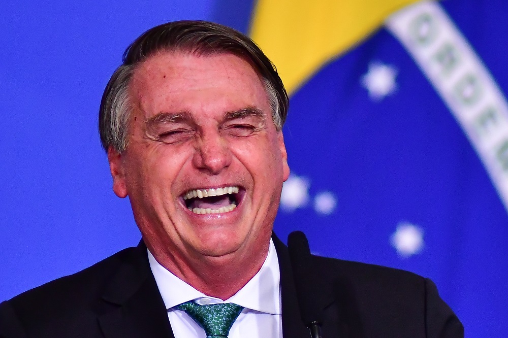 Jair Bolsonaro gargalha em frente a um fundo, aparentemente uma parede, com a bandeira do Brasil