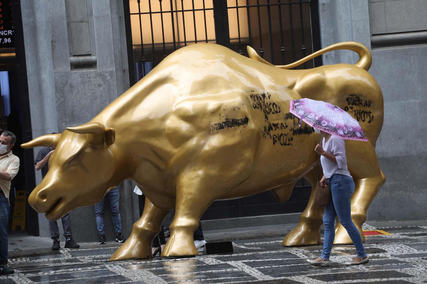 Transeunte com guarda-chuva passa em frente a estátua dourada de touro no centro de São Paulo