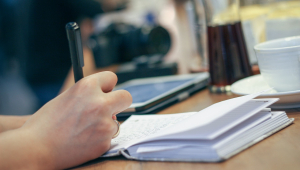 Uma mão segurando uma caneta e escrevendo em um caderno em uma meda com café e tablet