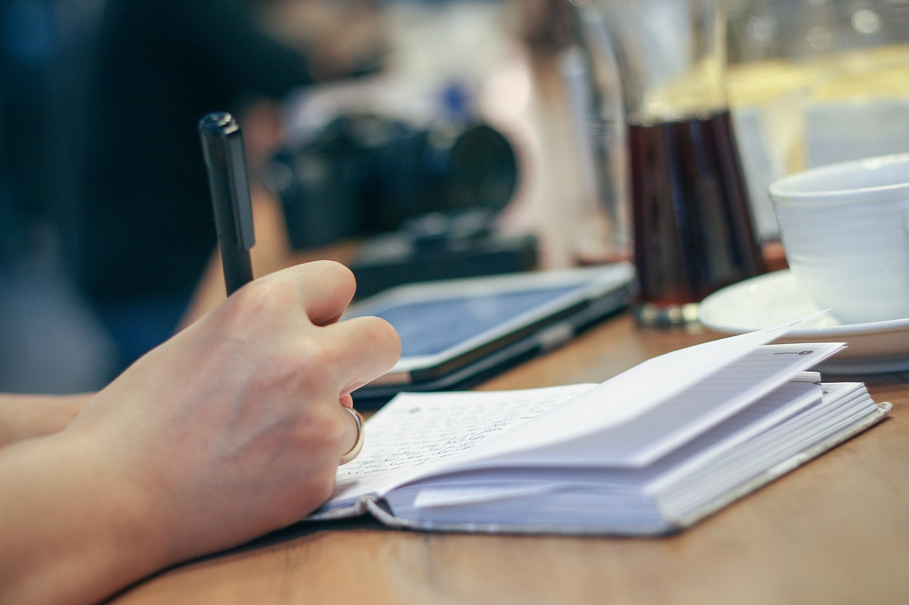 Uma mão segurando uma caneta e escrevendo em um caderno em uma meda com café e tablet