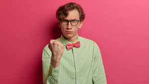 Jovem com visual nerd (branco, com cabelo loiro penteado para o lado, óculos de armação quadrada, camisa de linho verde e gravata borboleta rosa) à frente de um fundo rosa e expressando raiva e sentimento de vingança