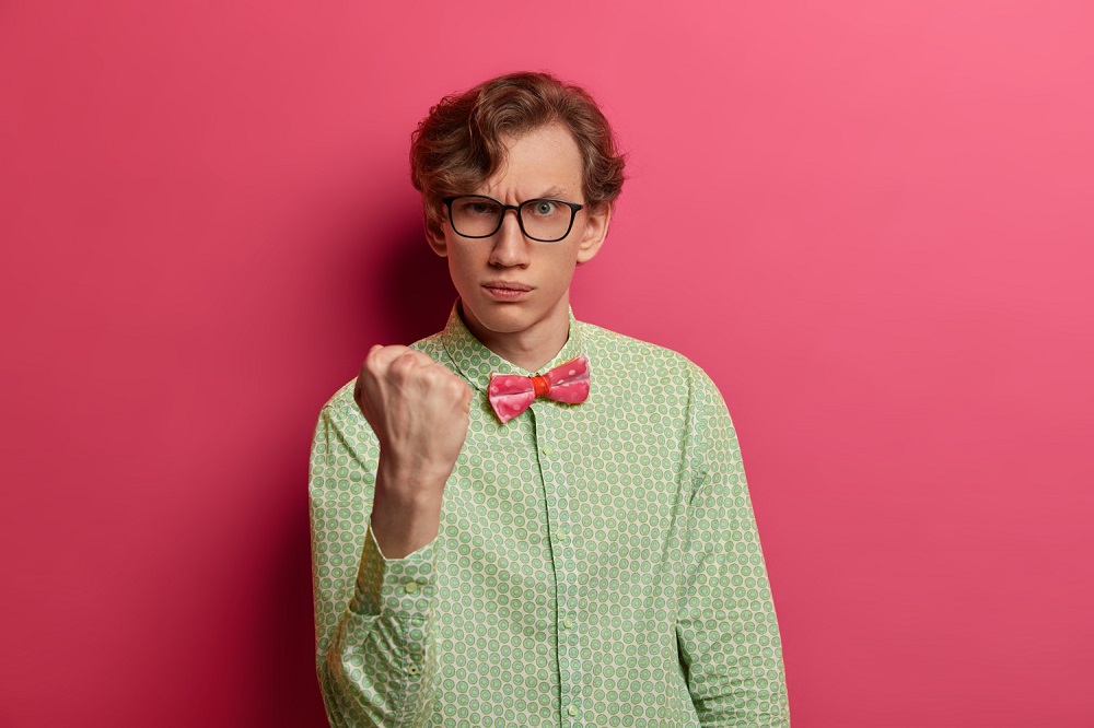 Jovem com visual nerd (branco, com cabelo loiro penteado para o lado, óculos de armação quadrada, camisa de linho verde e gravata borboleta rosa) à frente de um fundo rosa e expressando raiva e sentimento de vingança