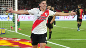 Atleta branco com camisa do River Plate (branca com faixa transversal vermelha) de braços abertos comemora após marcar gol