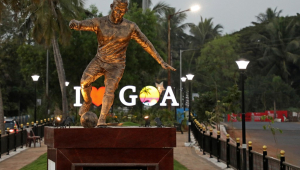 Estátua de bronze de jogador de futebol em frente a letreiro que diz 'eu amo Goa' em inglês