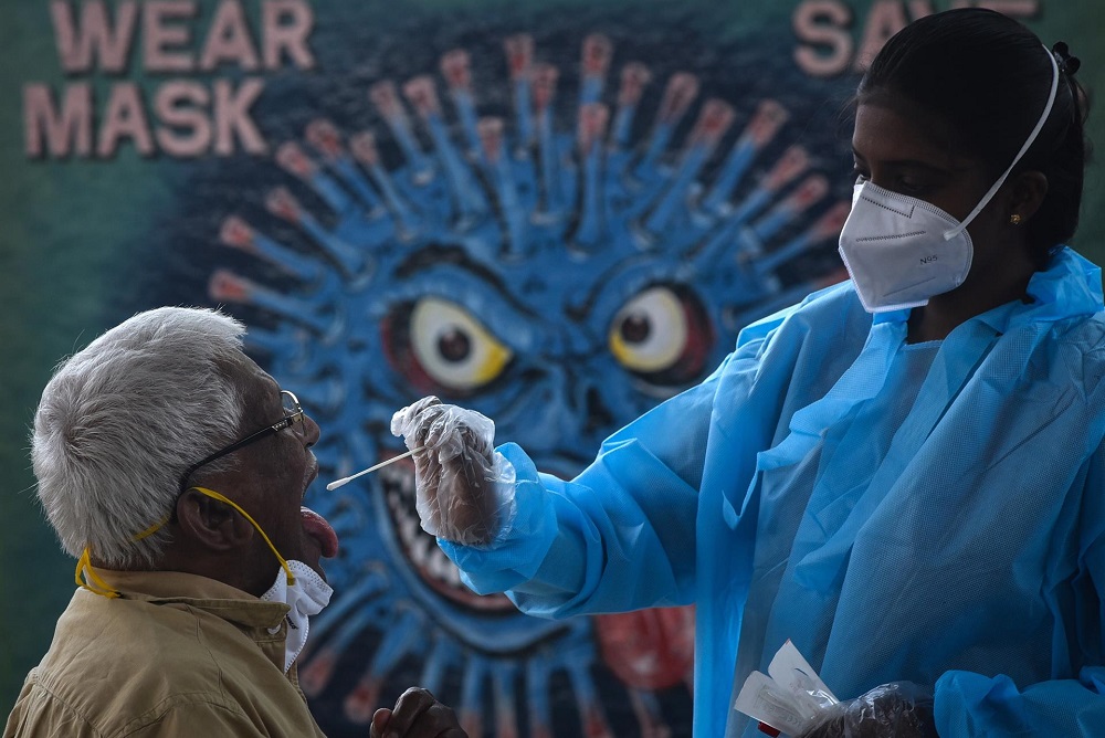 Agente de saúde da Índia coleta saliva de idoso para fazer teste de Covid-19, ao ar livre, em frente a um muro com o grafite de um vírus com cara maléfica