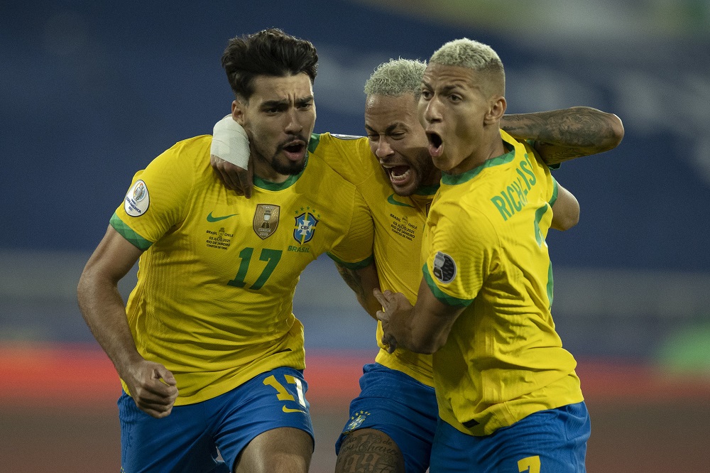 Paquetá, Neumar e Rocharlison correm abraçados, com cara de satisfação, comemorando gol da seleção brasileira; eles usam camisa amarela e calção azul