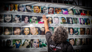 Familiar de vítima com camisa preta em frente a painel com as fotos das vítimas