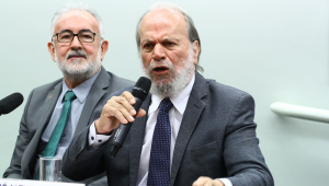 Dois homens brancos, de terno e gravata, sentados em uma sala na Câmara dos Deputados; o da direita tem barba longa e fala ao microfone