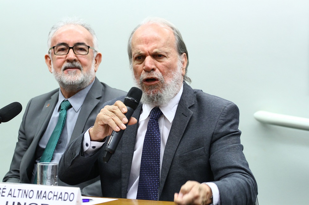 Dois homens brancos, de terno e gravata, sentados em uma sala na Câmara dos Deputados; o da direita tem barba longa e fala ao microfone