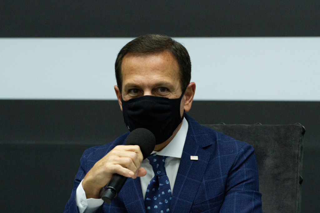 Governador João Doria: homem usando terno azul escuro e máscara preta segurando microfone