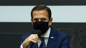 Governador João Doria: homem usando terno azul escuro e máscara preta segurando microfone