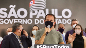 À frente de aliados e de um banner do governo do Estado de São Paulo, João Doria anuncia medidas em um púlpito, vestido de camiseta verde, com o microfone na mão direita