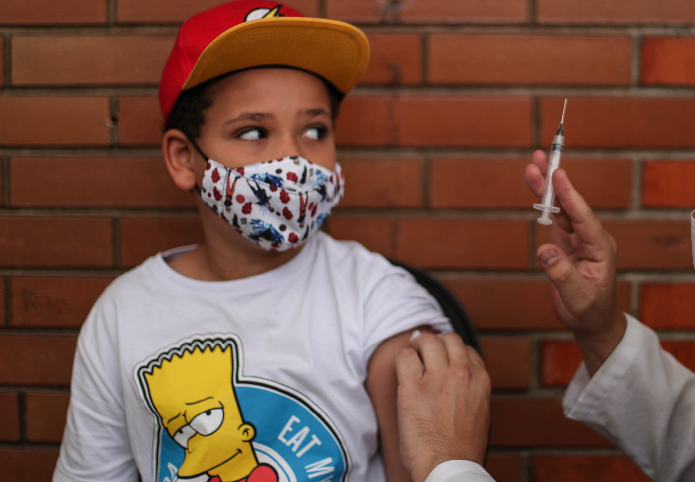 Menino com boné olhando para seringa com vacina contra a Covid-19 na mão da enfermeira