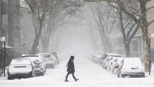 Pessoa bem agasalhada atravessa rua coberta por neve, com carros estacionados e igualmente cheios de neve