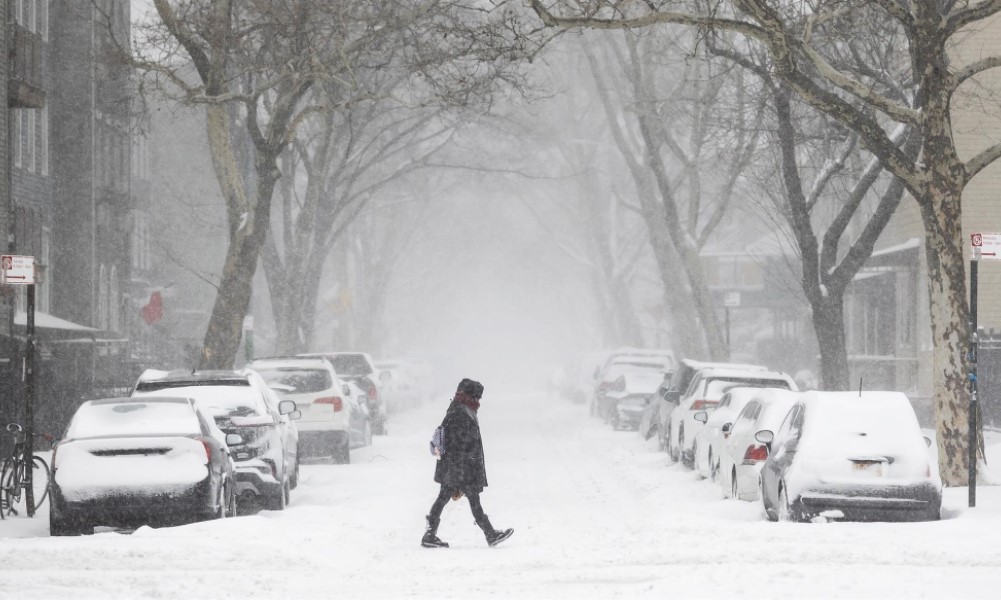 Pessoa bem agasalhada atravessa rua coberta por neve, com carros estacionados e igualmente cheios de neve