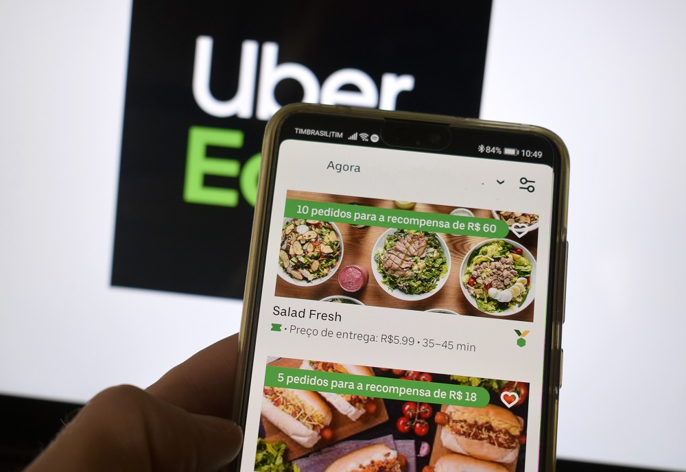Mão manuseia o celular logado no aplicativo Uber Eats (e o logo da empresa parece atrás)