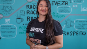 Mulher na faixa dos 30 anos , de cabelo preto, em frente a uma parede com desenhos e palavras que remetem ao empreendedorismo e à cultura de startups