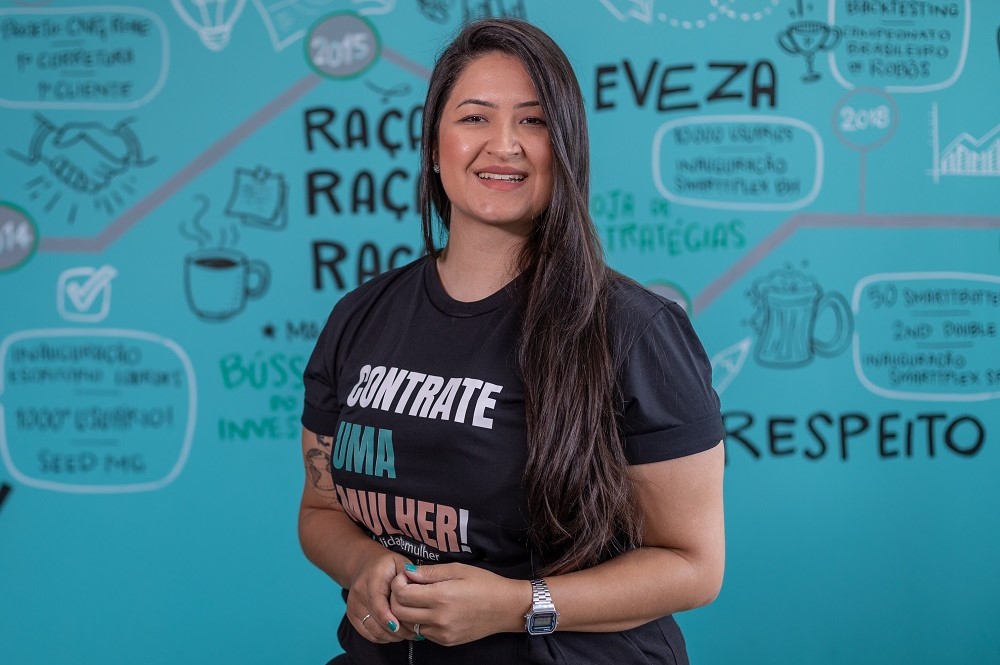 Mulher na faixa dos 30 anos , de cabelo preto, em frente a uma parede com desenhos e palavras que remetem ao empreendedorismo e à cultura de startups