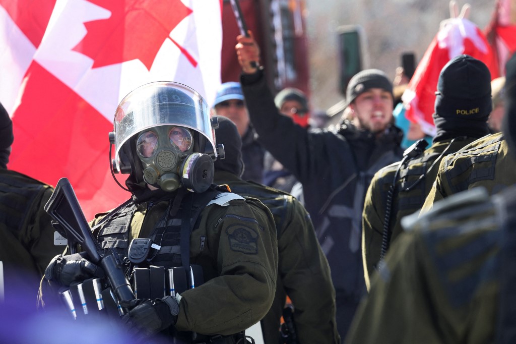 Policial com máscara de gás em frente a manifestante que segura a bandeia do Canadá