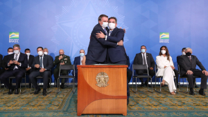 Em frente a um púlpito, Ciro Nogueira e Bolsonaro se abraçam; atrás deles, ministros e parlamentares assistem sentados à cena