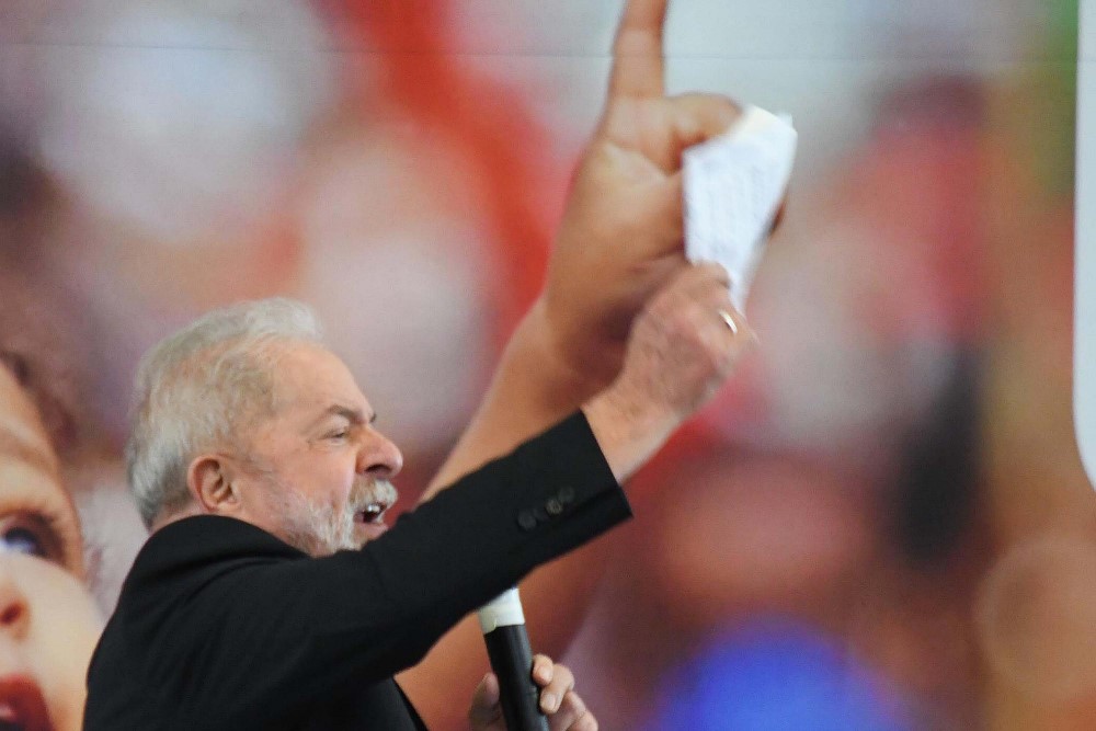 Em pé, de perfil, com os olhos cerrados e o braço direito levantado, Lula discursa ao lado de m banner que tem a imagem de uma criança, em tamanho gigante, com o braço igualmente levantado