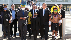 O presidente Jair Bolsonaro caminha ao lado de um, menino vestido de terno e gravata e de um indígena com roupas típicas; presidente da Funai está com um cocar