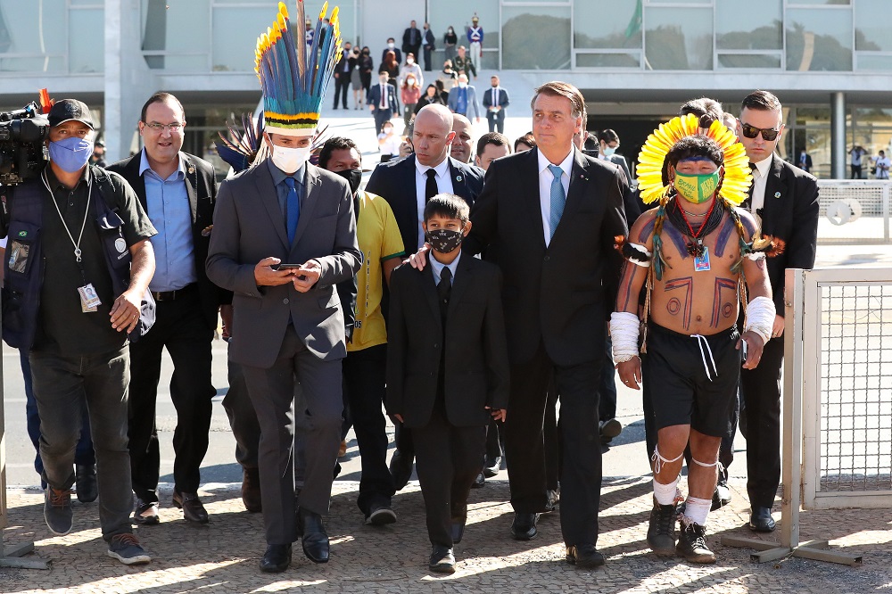 O presidente Jair Bolsonaro caminha ao lado de um, menino vestido de terno e gravata e de um indígena com roupas típicas; presidente da Funai está com um cocar
