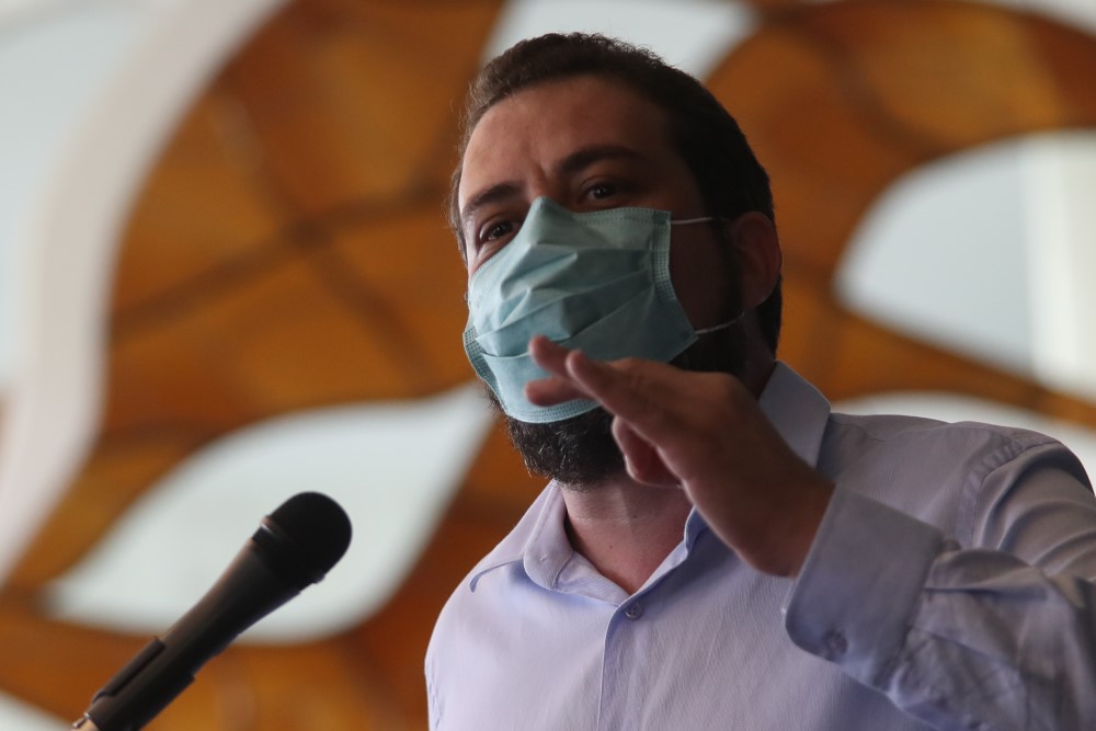 Guilherme Boulos, de máscara, gesticula com a mão esquerda enquanto fala diante de um microfone