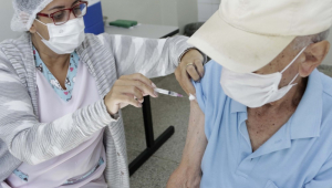 Enfermeira, com avental, máscara e touca, aplica a vacina no braço direito de um idoso de boné