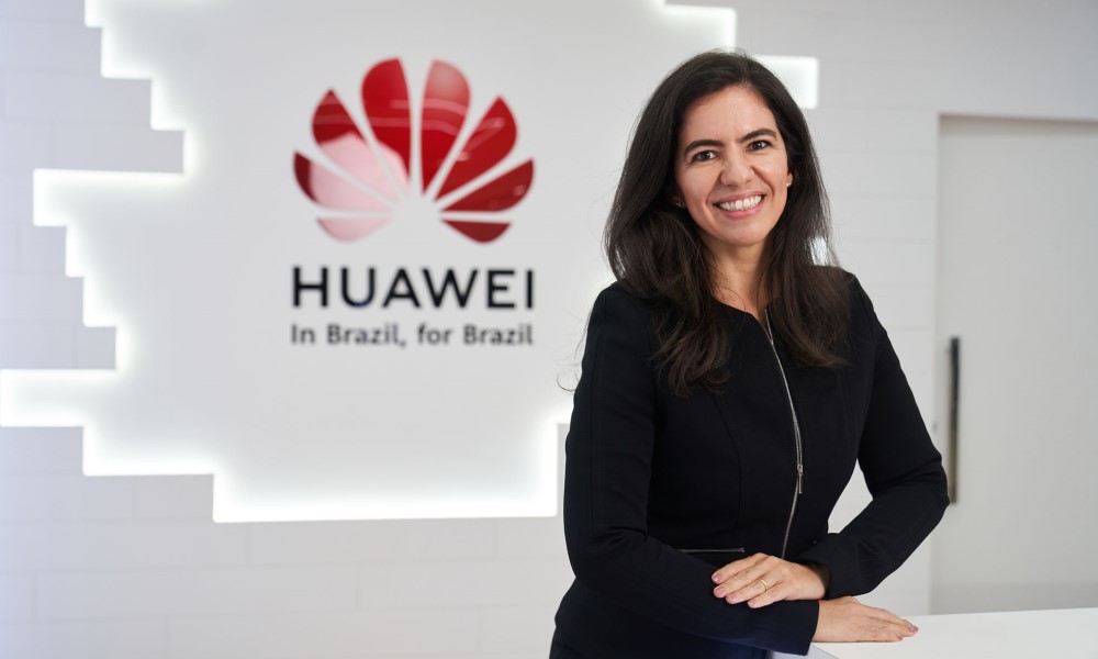 Em frente a um painel da Huawei, Ana Paula Barcelos de Sá (mulher entre 30/40 anos, branca e de cabelo preto), vestida de casaco preto, entrelaça os braços e sorri
