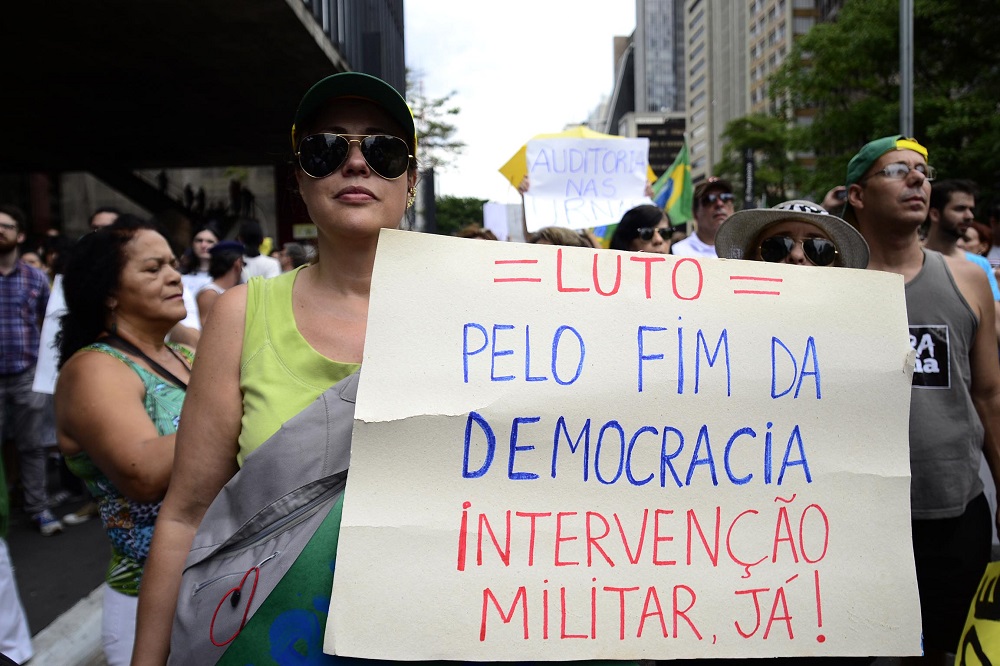 Durante manifestação, mulher de boné e óculos escuros segura cartaz em que diz que luta pelo fim da democracia e pede intervenção militar