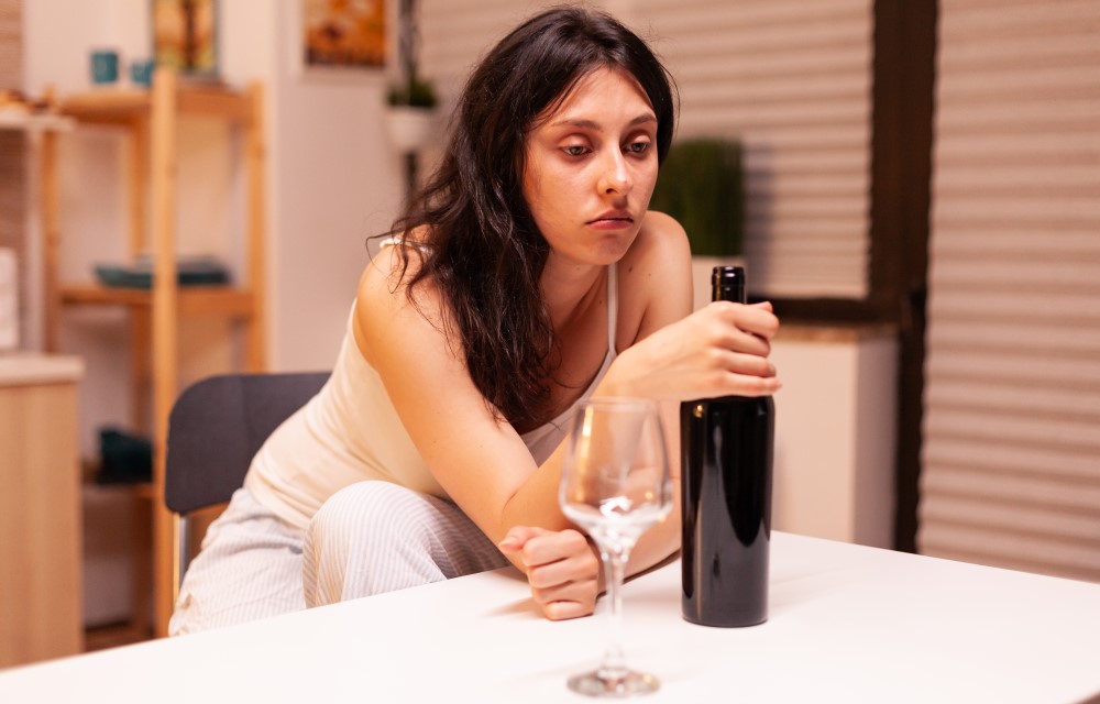 Jovem mulher branca e de cabelo preto segura uma garrafa de vinho sentada em frente a uma mesa em casa