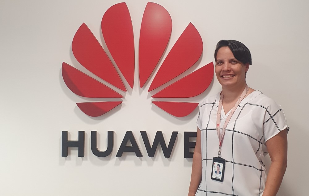 Mulher branca na faixa dos 30/40 anos posa com crachá na frente do símbolo da Huawei