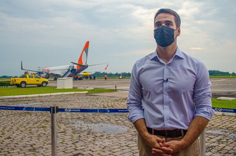 Eduardo Leite, de camisa,, calça social e máscara, se posiciona à frente de uma pista de pouso, com um avião estacionado