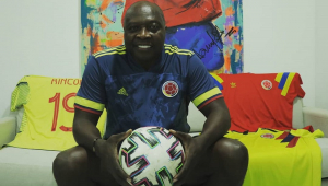 O ex-jogador Rincón sentado em um sofá amarelo, com a camisa azul da Colômbia, calça jeans e segurando uma bola de futebol