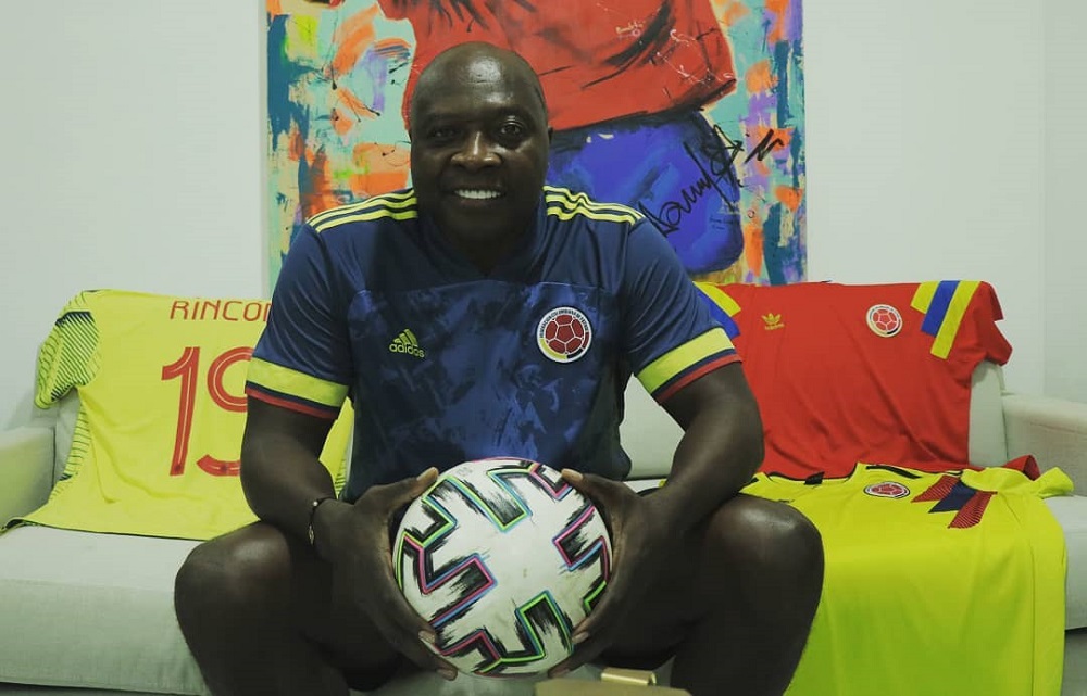 O ex-jogador Rincón sentado em um sofá amarelo, com a camisa azul da Colômbia, calça jeans e segurando uma bola de futebol