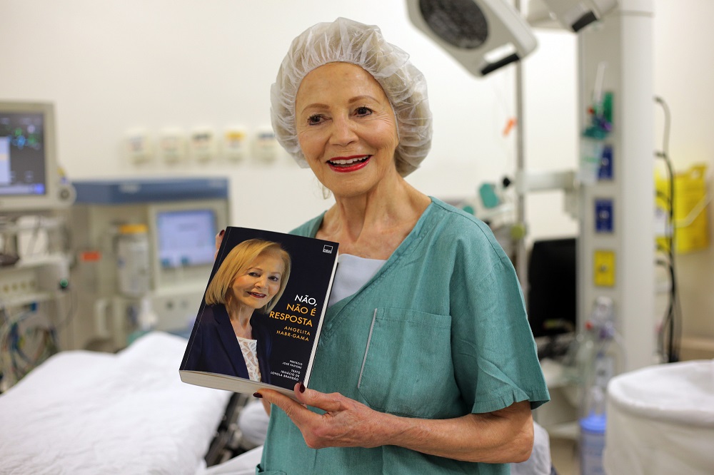A médica Angeluta Gama, de 80 anos, vestida com toupa de cirrugiã, segura o seu livro autobiográfico