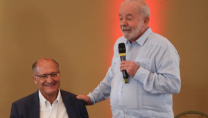 Lula fala ao microfone coloca a mão direita no ombro de Geraldo Alckmin, que está sentado