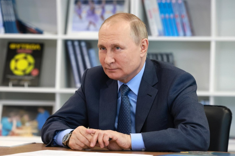 Vladimir Putin, trajado com terno azul marinho e camisa azul clara e gravata, sentado em uma cadeira em frenta a uma estante com livros