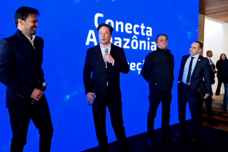 Elon Musk discursa ao lado de Jair Bolsonaro e Fabio Faria em frente a um painel onde está escrito Conecta Amazônia