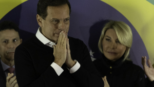 Ao lado da mulher, Bia Doria, à frente de uma bandeira do Brasil, João Doria faz sinal de reza com as mãos