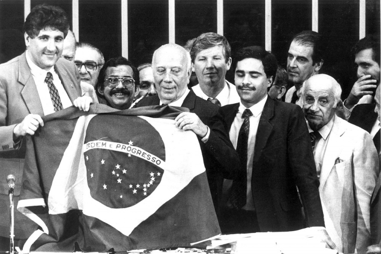 Ao lado de vários parlamentares, o presidente da Câmara dos Deputados, Ulisses Guimarães, segura a bandeira do Brasil na última sessão da Assembléia Constituinte