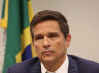 Roberto Campos Neto com expressão séria e bandeira do Brasil ao fundo