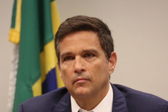 Roberto Campos Neto com expressão séria e bandeira do Brasil ao fundo