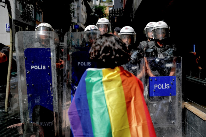 Manifestante aparentemente do sexo feminino, com bandeira do arco-íris amarrada nas costas, encara polícais em rua de Istambul
