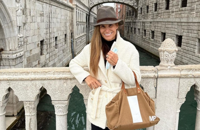 Mariana Papa, uma mulher na faixa dos 40 anos, posa agasalhada e usando bolsa e chapéu em uma ponte em Veneza