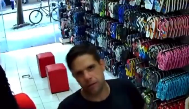Homem branco de camiseta preta olha para câmera de segurança de uma loja de calçados