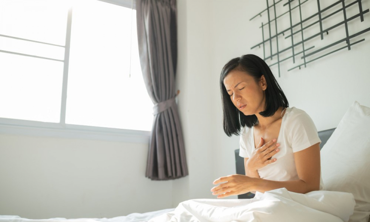 Mulher asiática sentada na cama coloca a mão no peito e aparenta ter ataque cardíaco