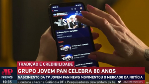 Frame de reportagem sobre os 80 anos da Jovem Pan mostra mão segurando um celular em que vídeo do Pingos nos Is é exibido